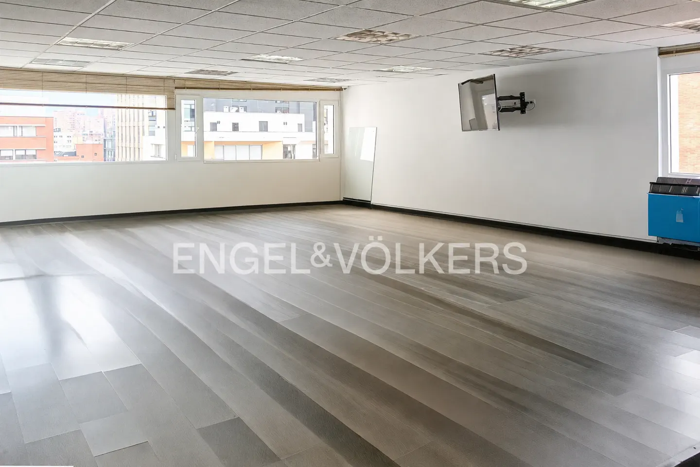 Bright, empty office space with gray wood floors, white walls, and large windows. A TV is mounted on the wall.