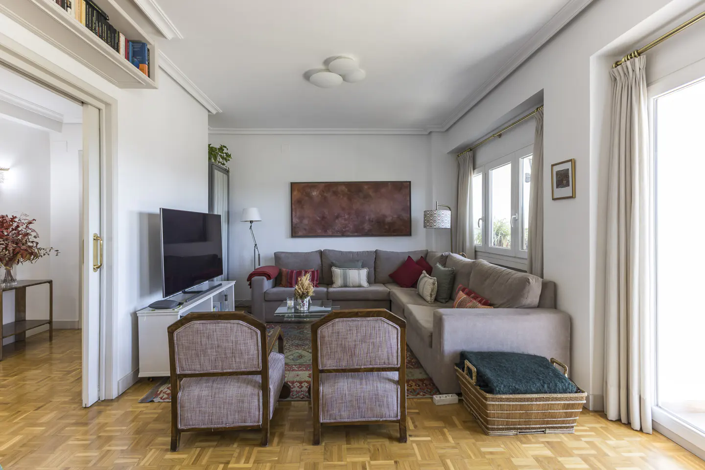 Living room with parquet floors, a gray sectional sofa with colorful pillows, and two armchairs. A TV sits on a white console.