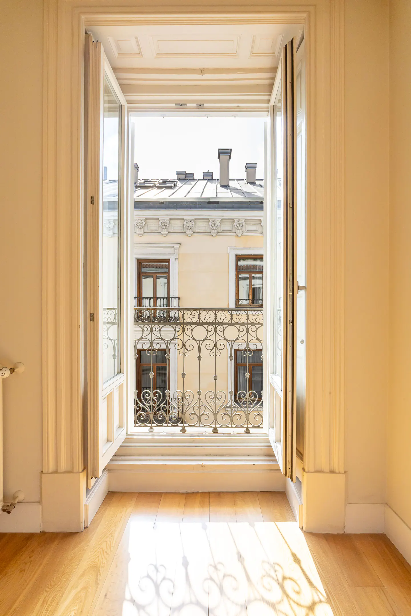 Open white window with curtains, overlooking a building with a wrought iron balcony and brown windows. Light wood floor.