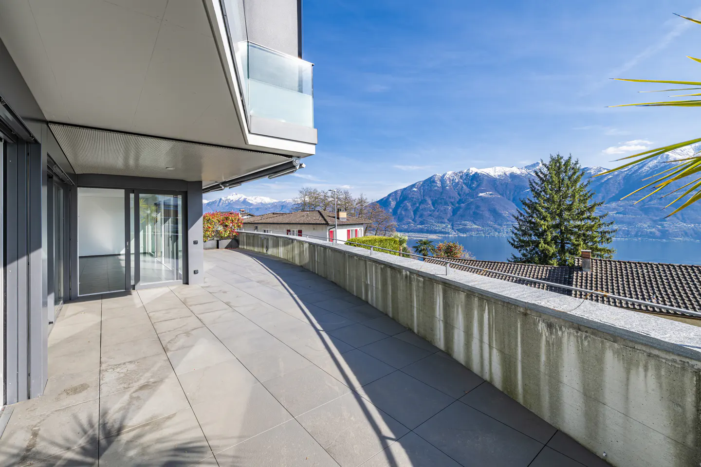 Balcony view of lake and mountains. Gray stone patio with glass railing. Blue sky.