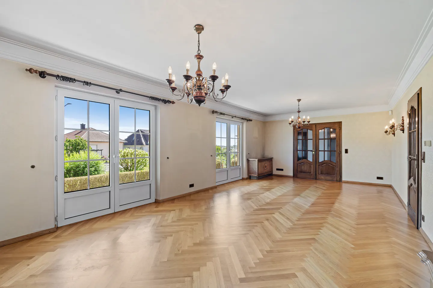 Spacious, empty room with light wood herringbone floors, two ornate chandeliers, and glass doors leading to a garden.