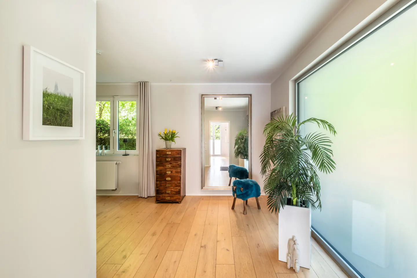 Bright hallway with wood floors, a large mirror, and a potted palm tree by a frosted window. A wooden dresser sits near a window with yellow flowers.