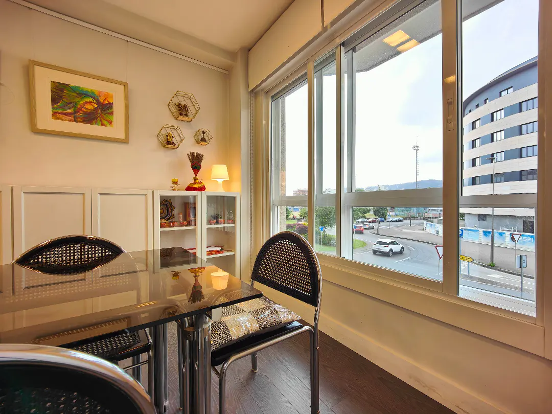 A bright dining room with a glass table, wicker chairs, and a large window overlooking a city street.