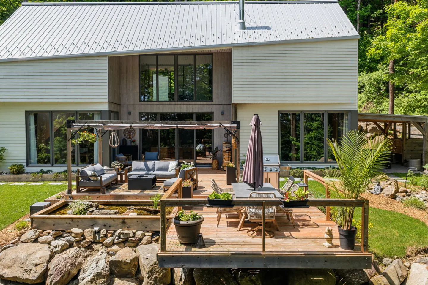 Modern two-story home with a large wooden deck, outdoor seating, dining area, and a small pond, set in a wooded landscape.