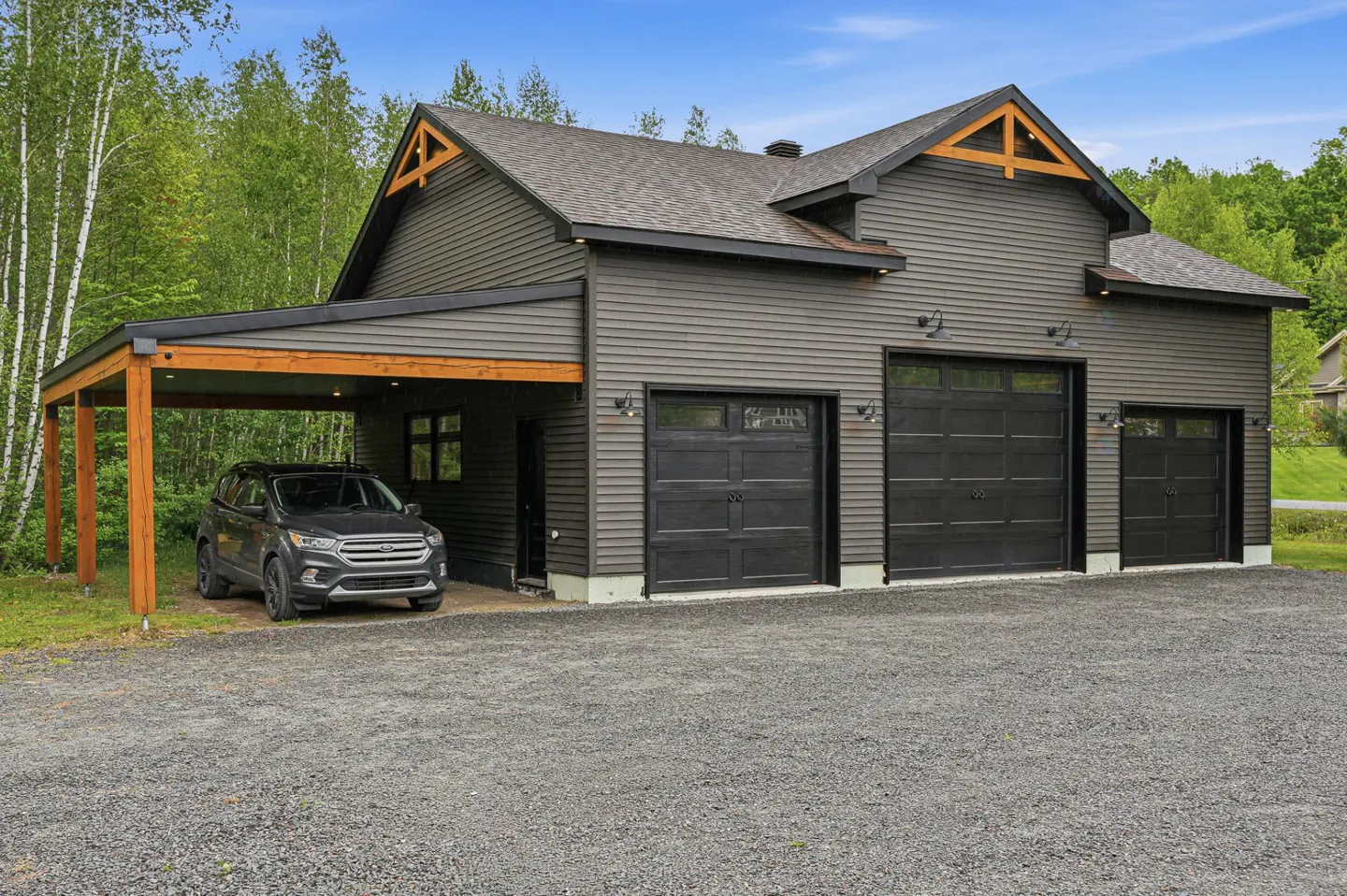 Dark gray garage with three black doors and a wooden carport sheltering a gray SUV on a gravel driveway near trees.