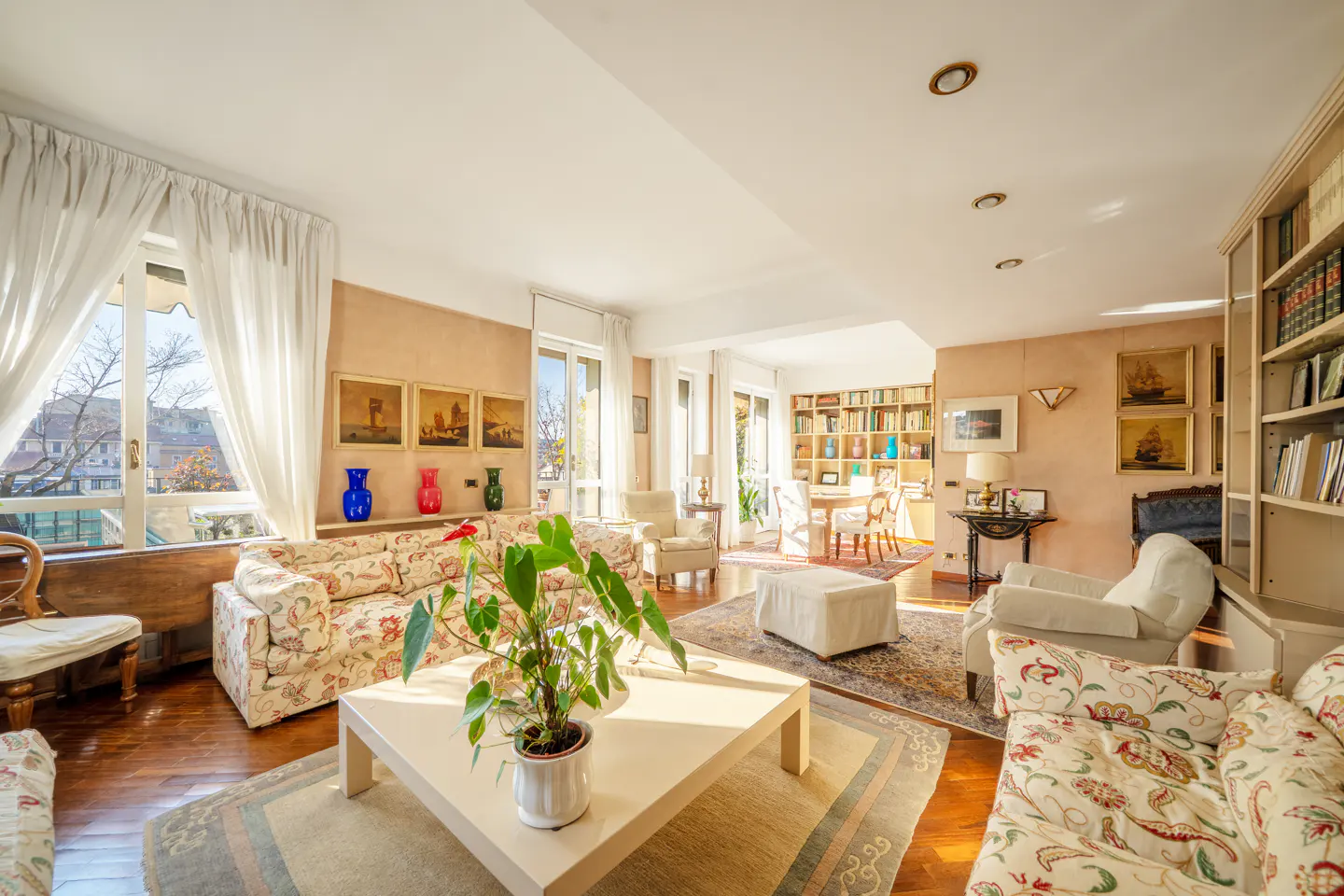 Bright living room with floral sofas, white coffee table with plant, and large windows with white curtains. Bookshelves and artwork add character.