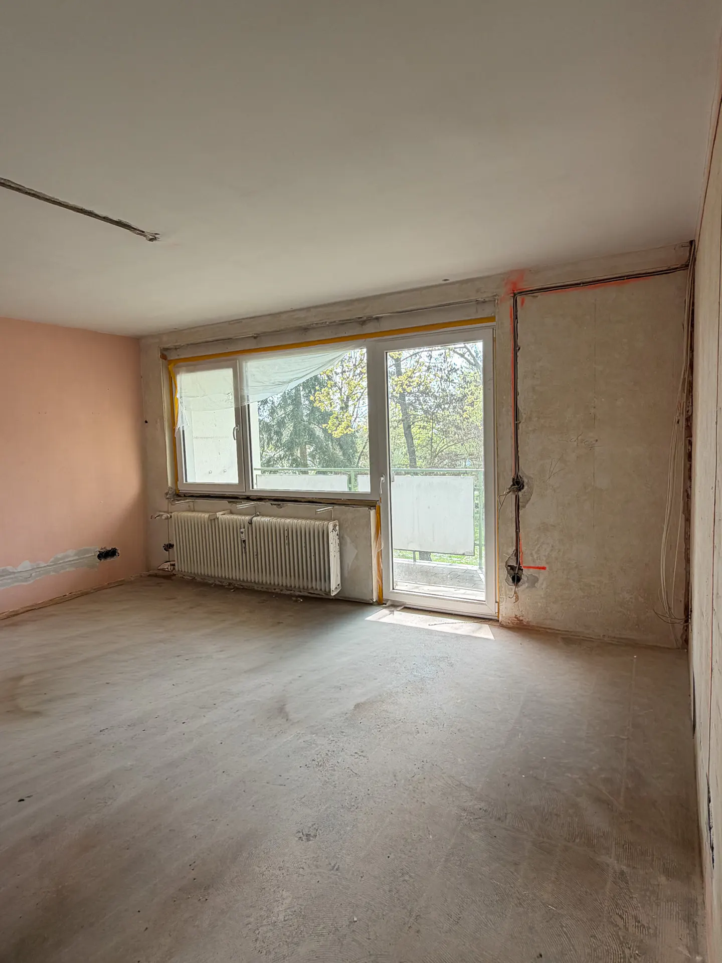 Empty room with pink wall, concrete floor, radiator, and window with trees outside. Electrical wiring is exposed on the wall.