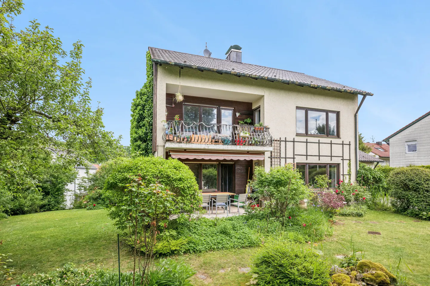 Two-story house with a balcony, patio furniture, and a lush green garden under a clear blue sky.