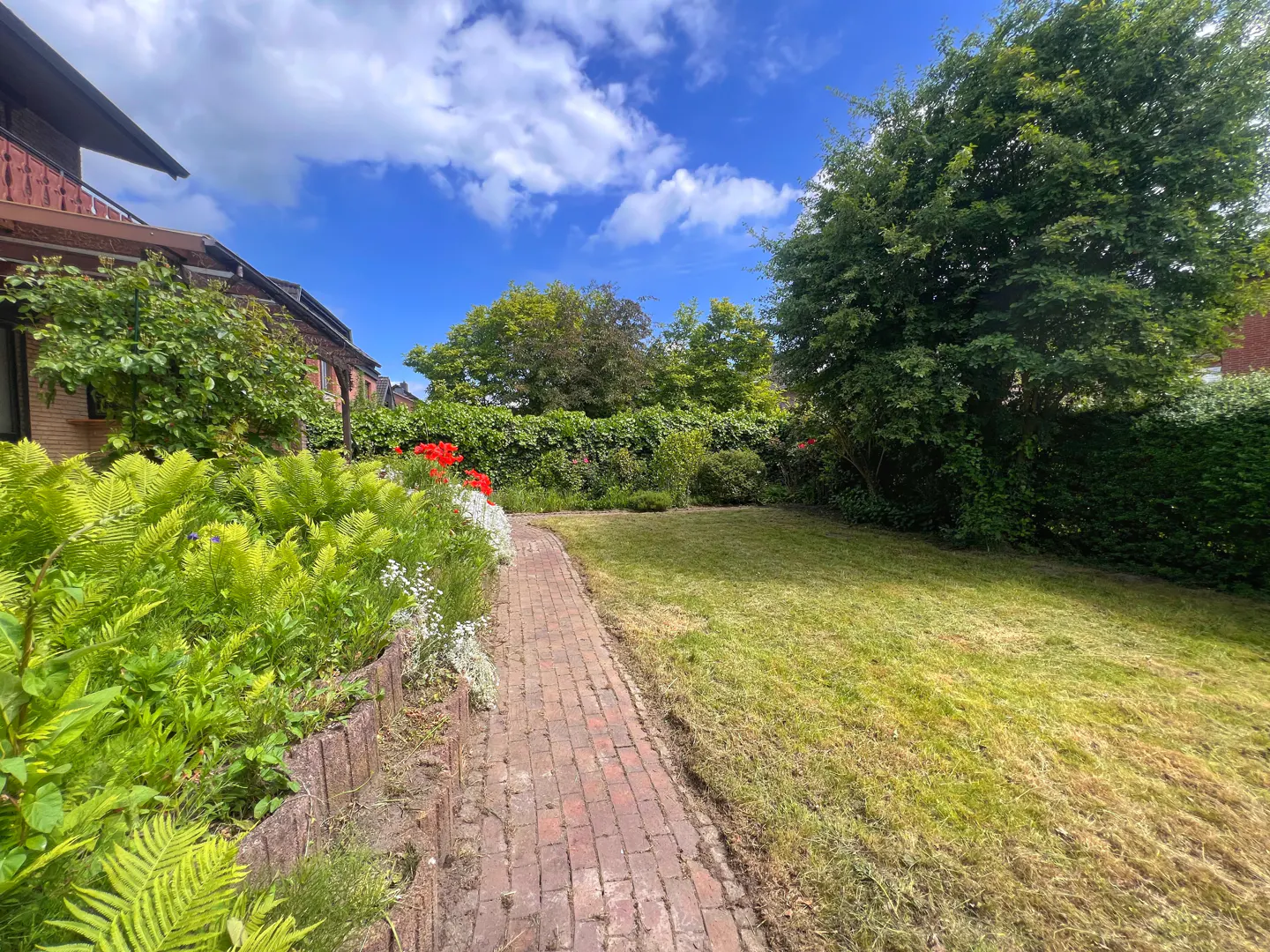 Brick path leads through a lush green lawn and flower garden, under a blue sky with fluffy white clouds.