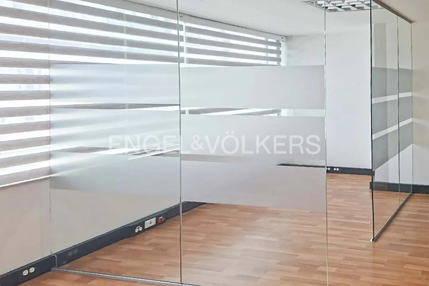 Office interior with glass walls, wood floor, and horizontal blinds. The Engel & Völkers logo is on the glass.