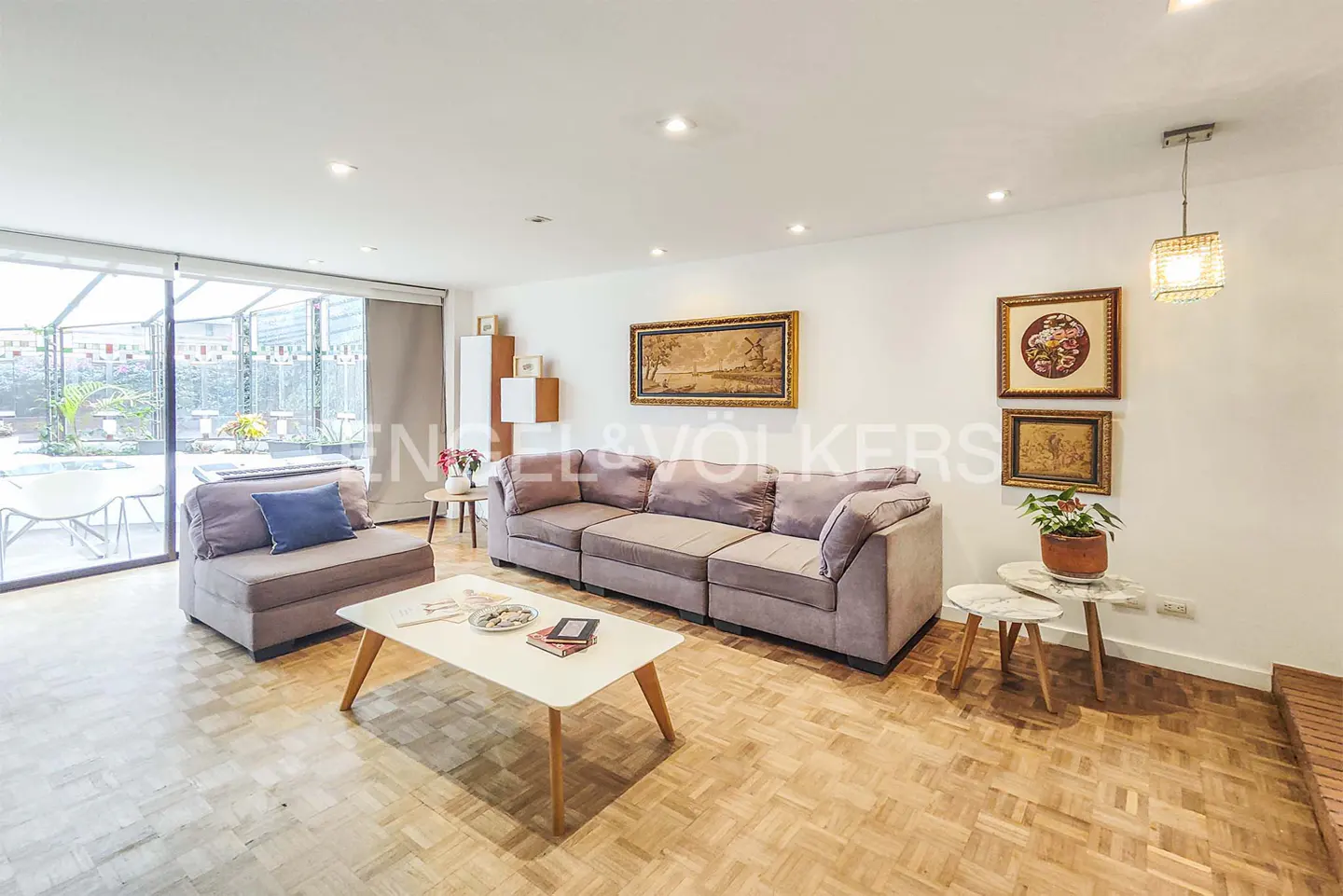 Living room with a gray sofa, white coffee table, and parquet floors. Artwork hangs on the white walls. A sunroom is visible in the background.