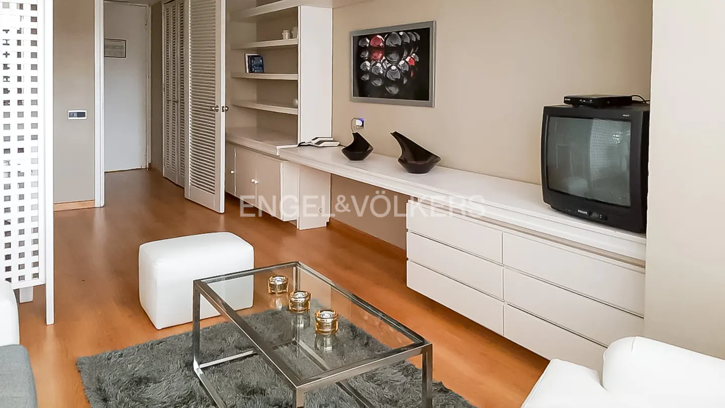 Modern living room with a glass coffee table, white ottoman, long white desk, and a vintage television on a white cabinet.