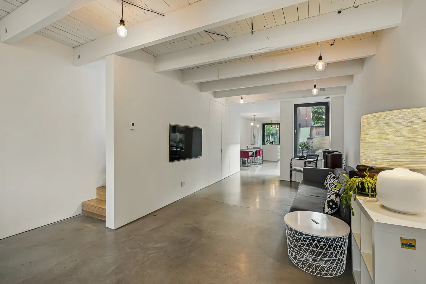 Bright, modern living room with white walls, concrete floors, and exposed beams. A gray sofa and white furniture add to the minimalist aesthetic.