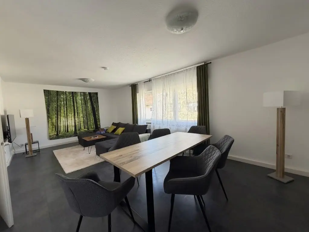 Bright living room with a wood table, gray chairs, and a gray sectional sofa with yellow pillows. A forest tapestry hangs on the wall.