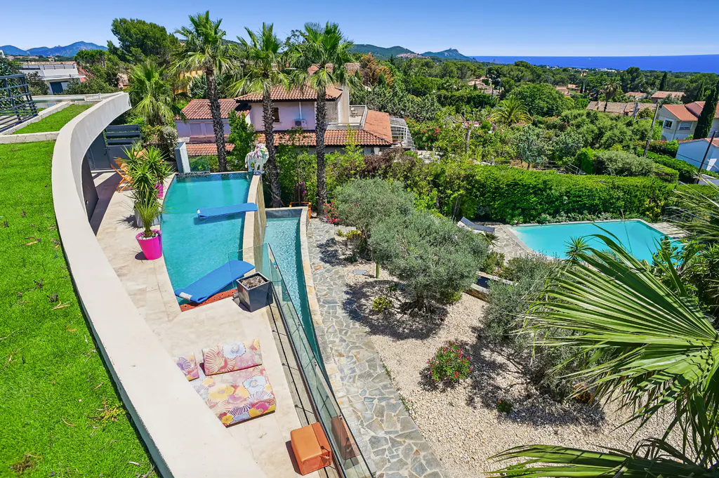 High-angle view of a luxury villa with two swimming pools, palm trees, a stone patio, and a distant view of the blue sea.