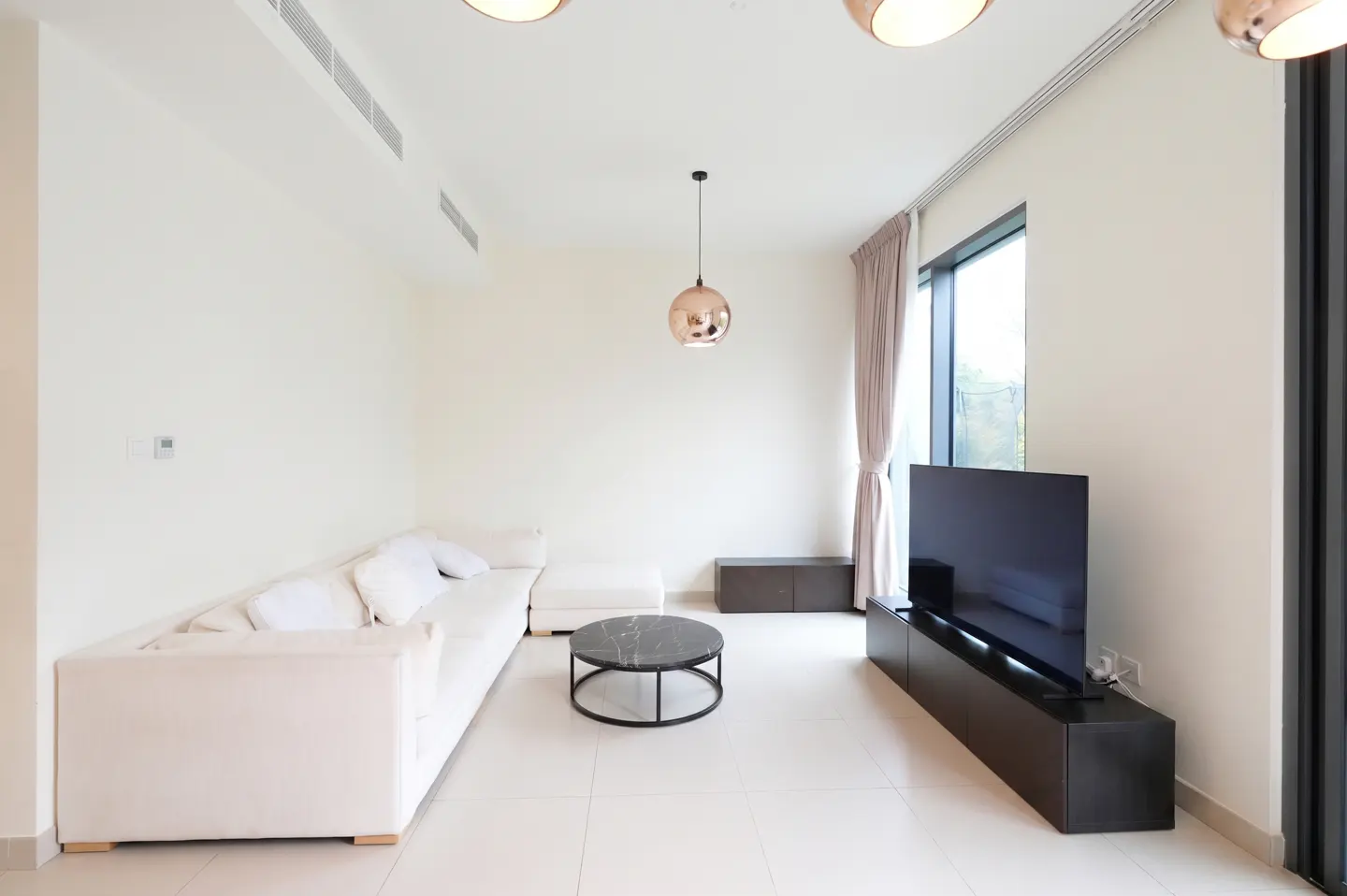 Bright living room with white walls, tile floor, and sectional sofa. A black TV and round table add contrast.