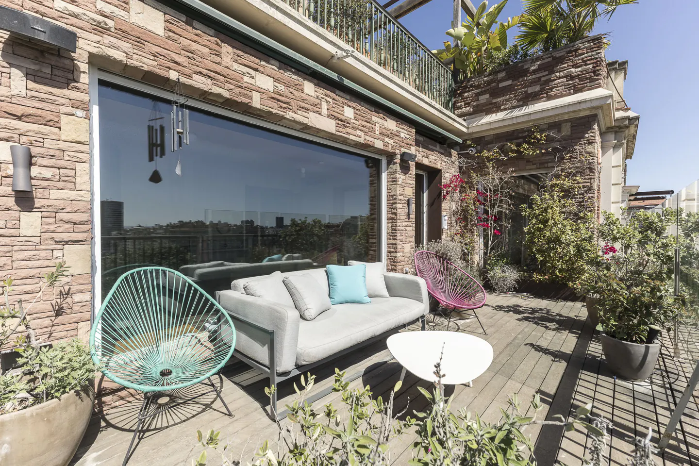 A rooftop patio with a gray sofa, blue pillow, teal and pink chairs, and a white table. The patio has brick walls and plants.