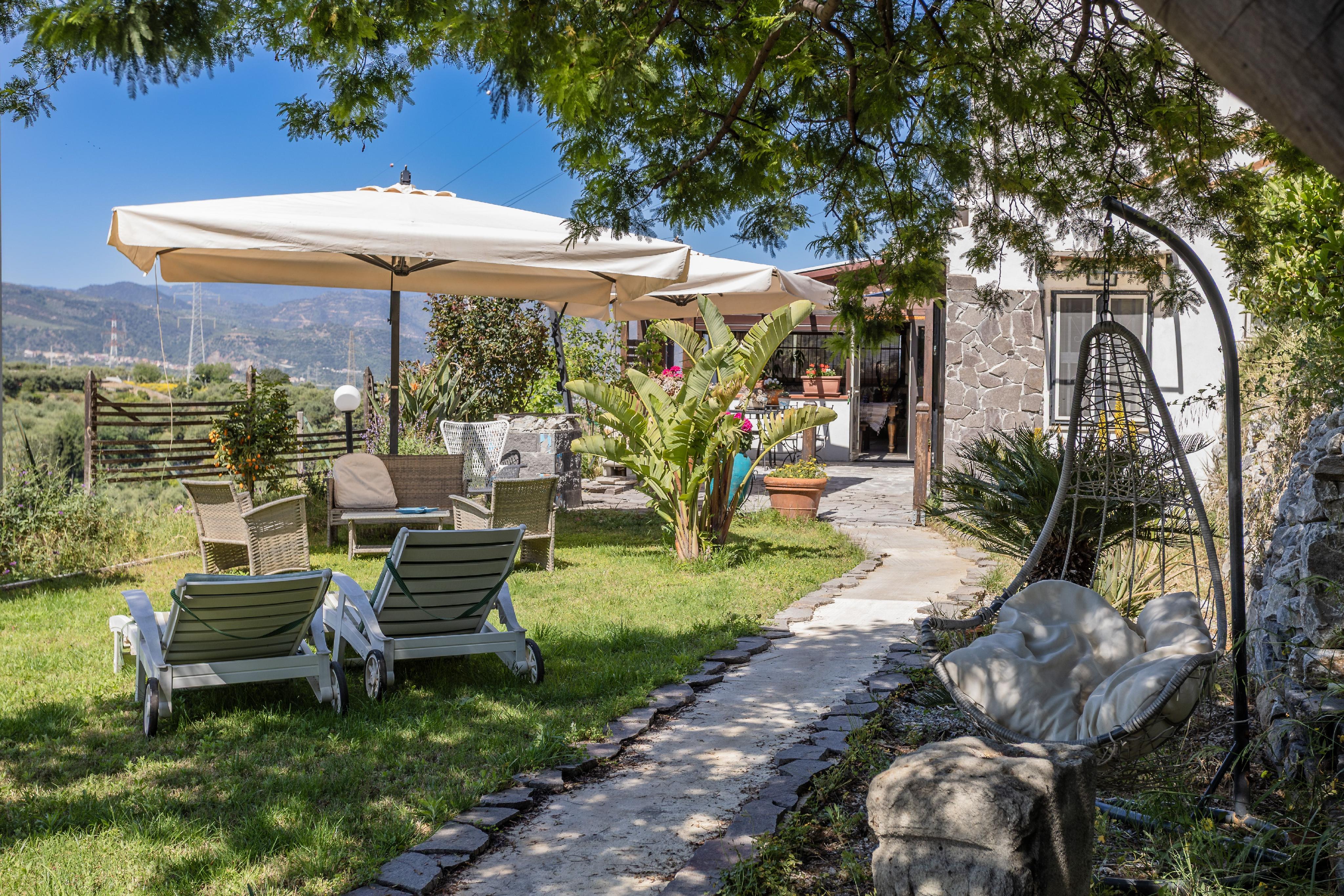 Sunny garden patio with lounge chairs, a hanging egg chair, and large umbrellas, overlooking mountains under a blue sky. [AI-generated text]