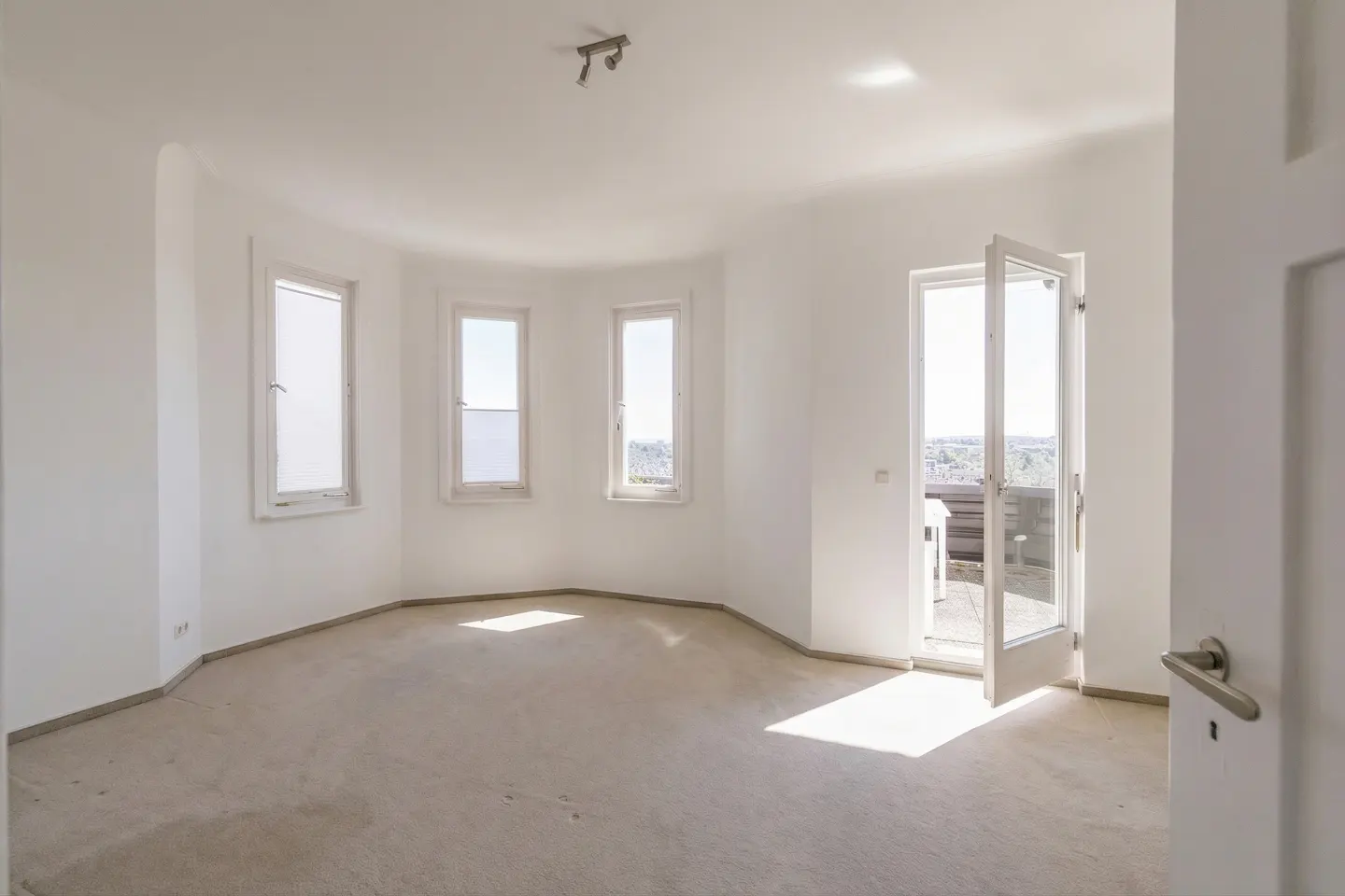 Bright, empty room with white walls, beige carpet, and three windows. Open glass doors lead to a balcony with a view.