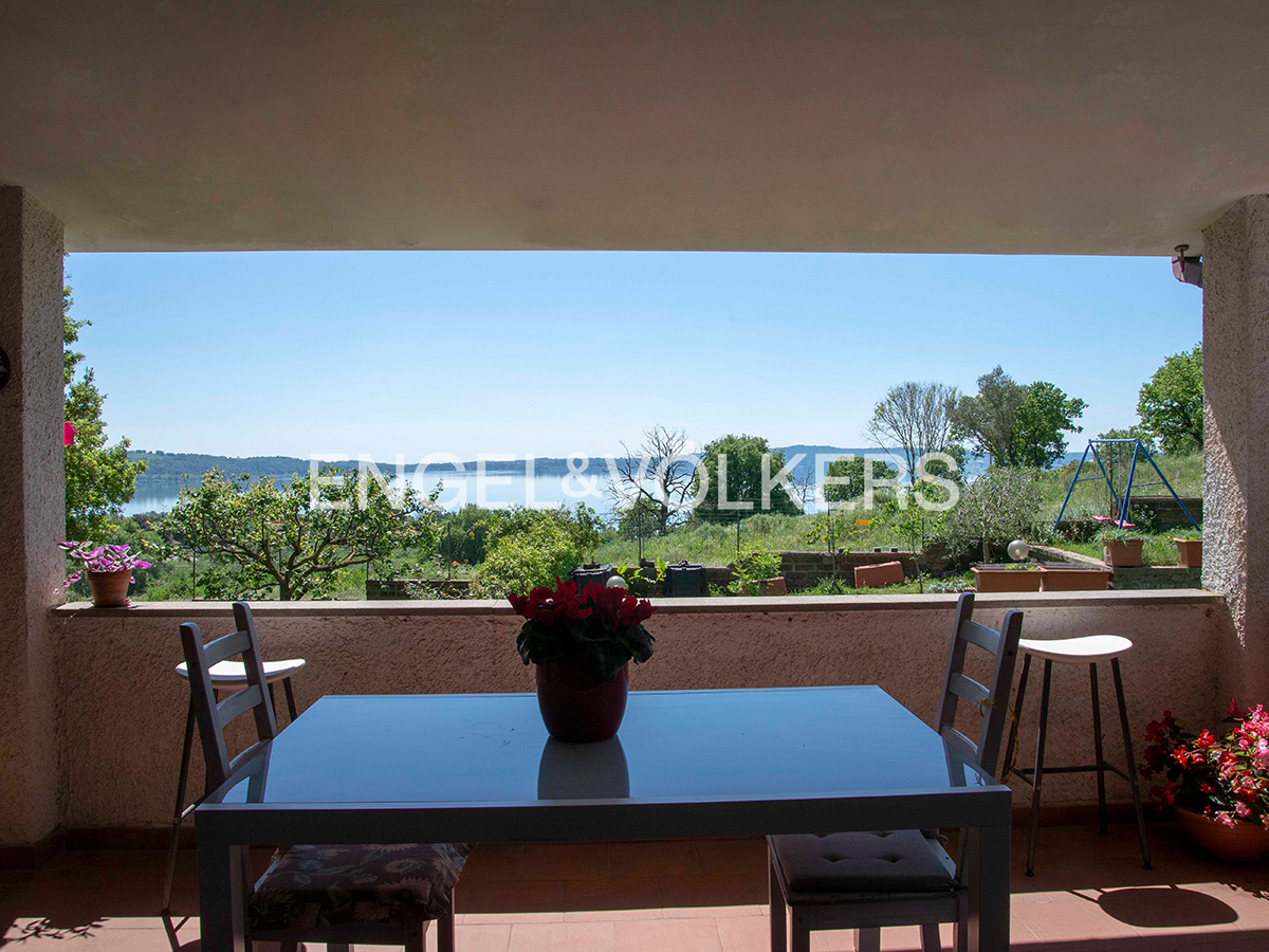 Covered patio with table, chairs, and flowers overlooking a lake and green landscape on a sunny day.