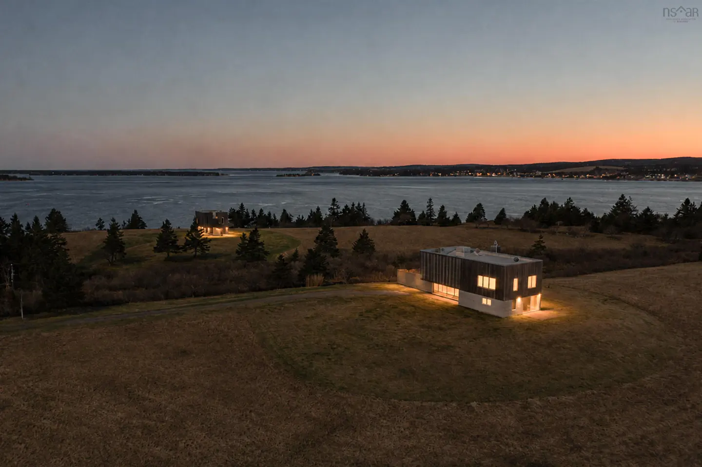 Distant aerial view of two modern homes on a grassy hill overlooking a bay at sunset.