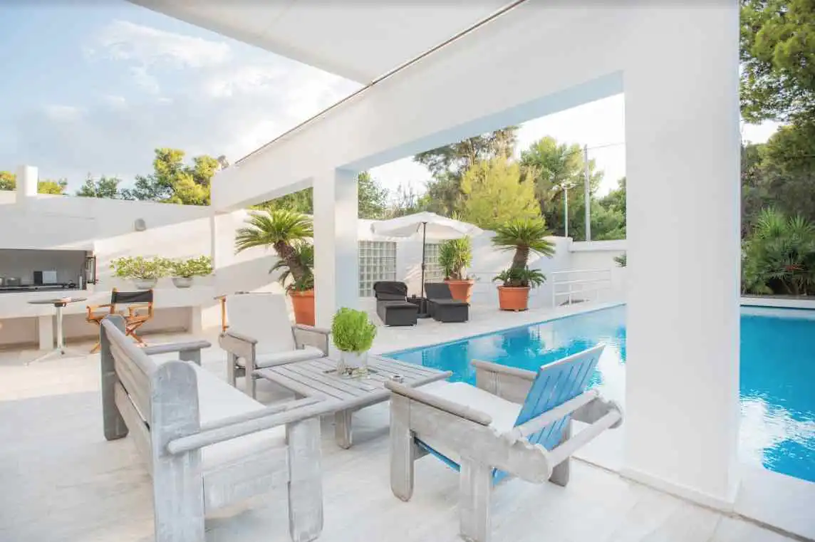 Outdoor patio with gray wooden furniture, a blue pool, and white walls under a bright sky.