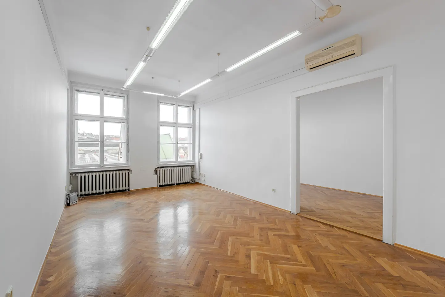 Bright, empty room with white walls, herringbone wood floor, two windows with radiators, and an open doorway.