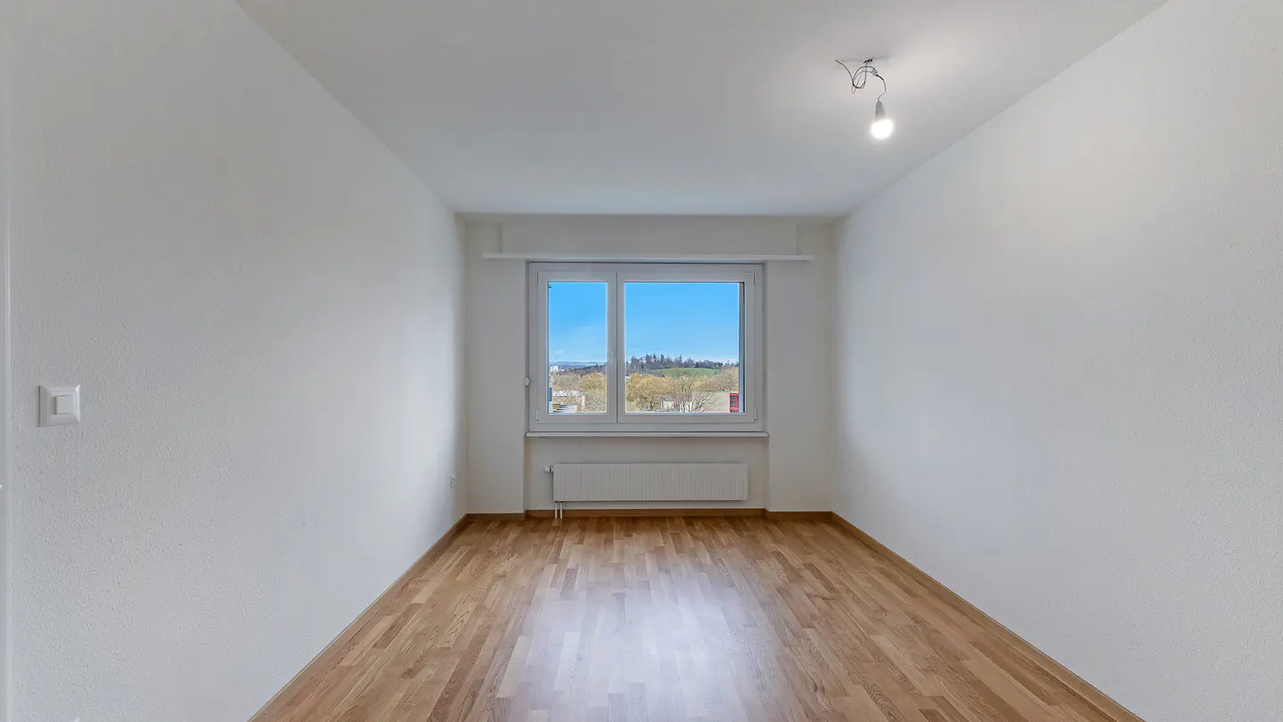 Empty room with hardwood floors and white walls. A window shows a view of trees and a blue sky.
