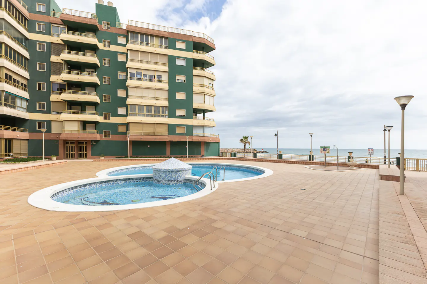 Exterior view of a green apartment building with a blue tiled pool and ocean view.