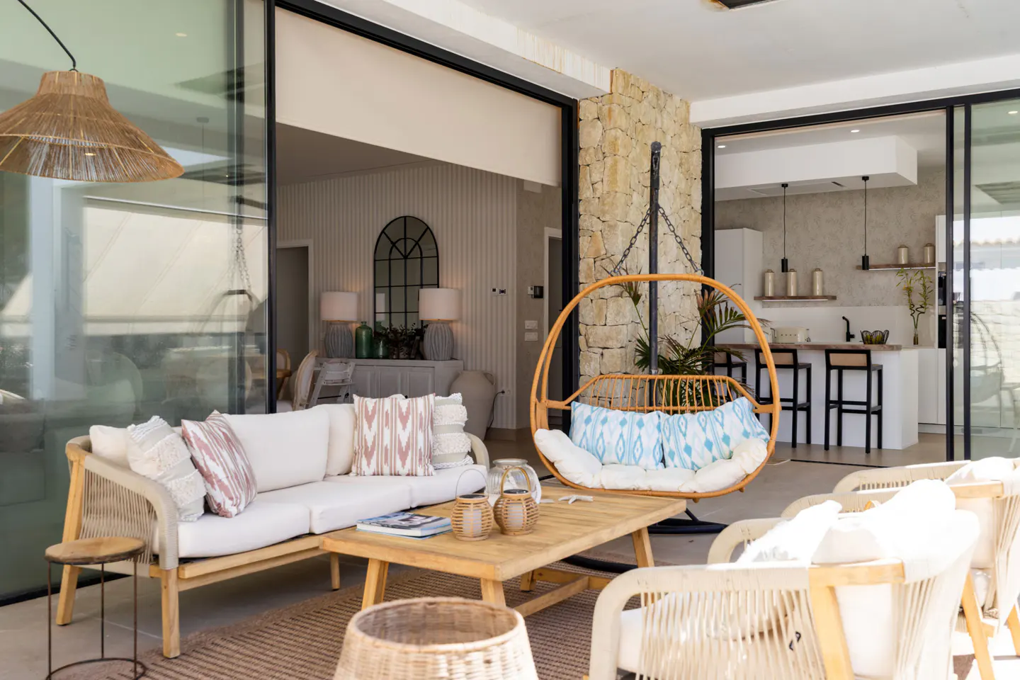A bright patio with a sofa, chairs, and a hanging chair. Sliding glass doors lead to the interior of the house, including a kitchen and living room.