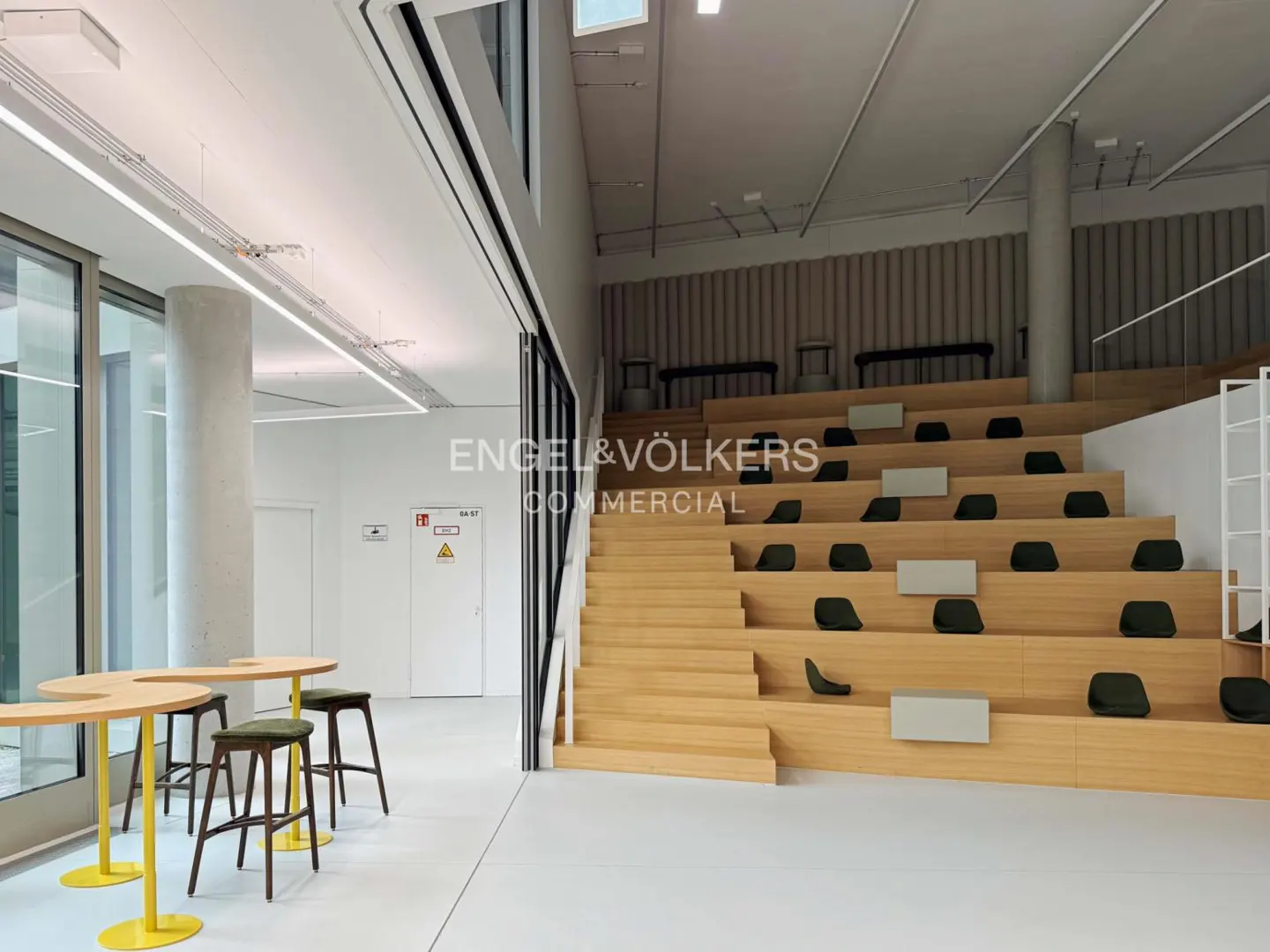 Bright, modern office space with tiered seating, yellow tables, and stools. The room is spacious and well-lit.