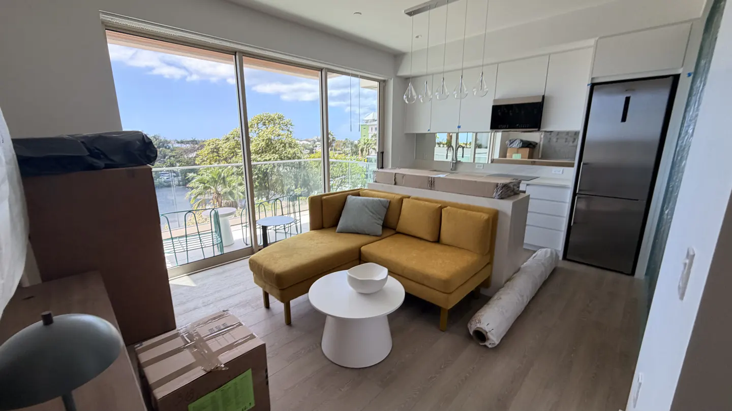 Bright apartment interior with a yellow sectional sofa, white kitchen cabinets, and a stainless steel refrigerator. Boxes are scattered around the room. Balcony view.