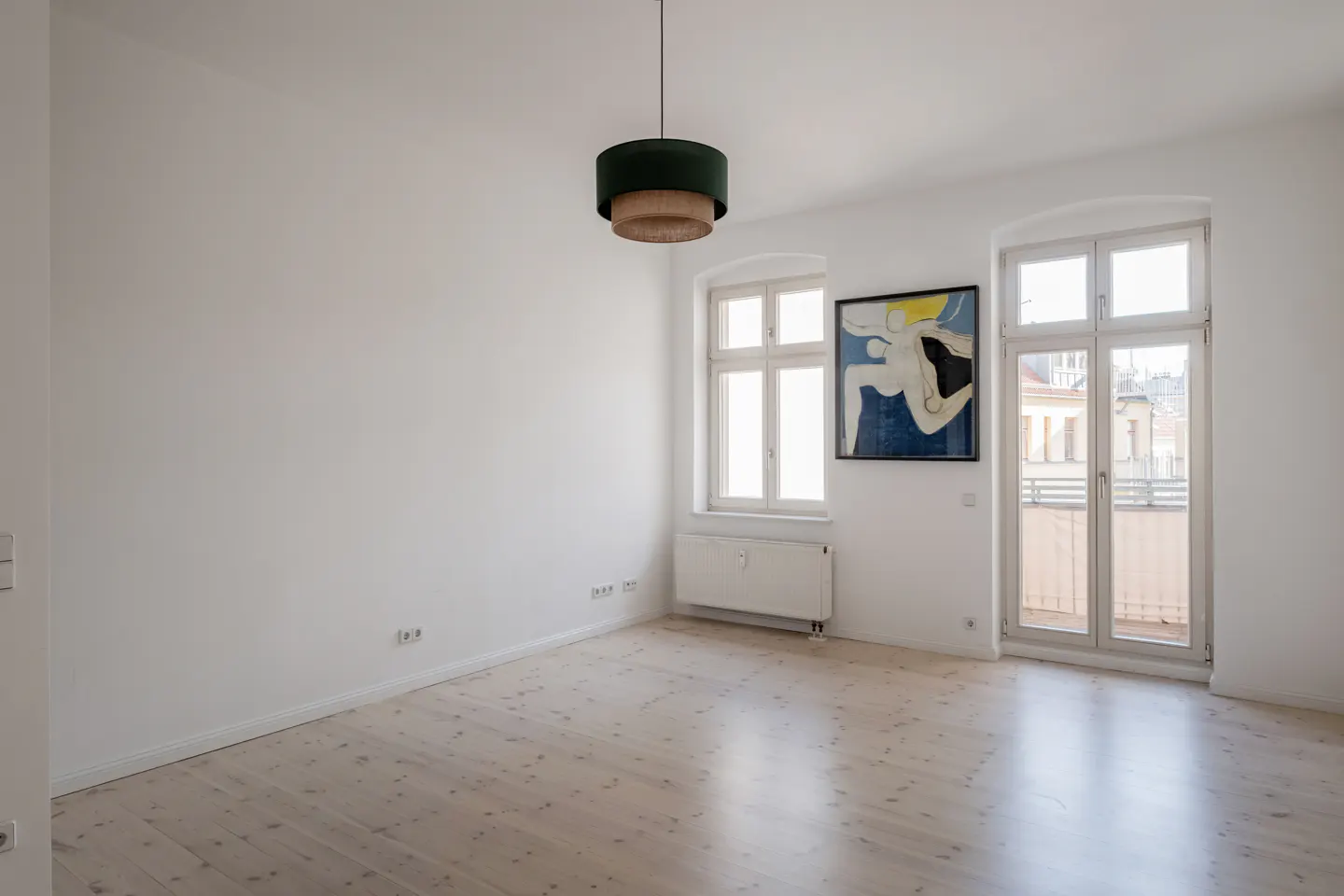 Bright, empty room with white walls, light wood floors, and two windows. A modern, two-tiered green and tan lamp hangs from the ceiling. An abstract painting is on the wall.