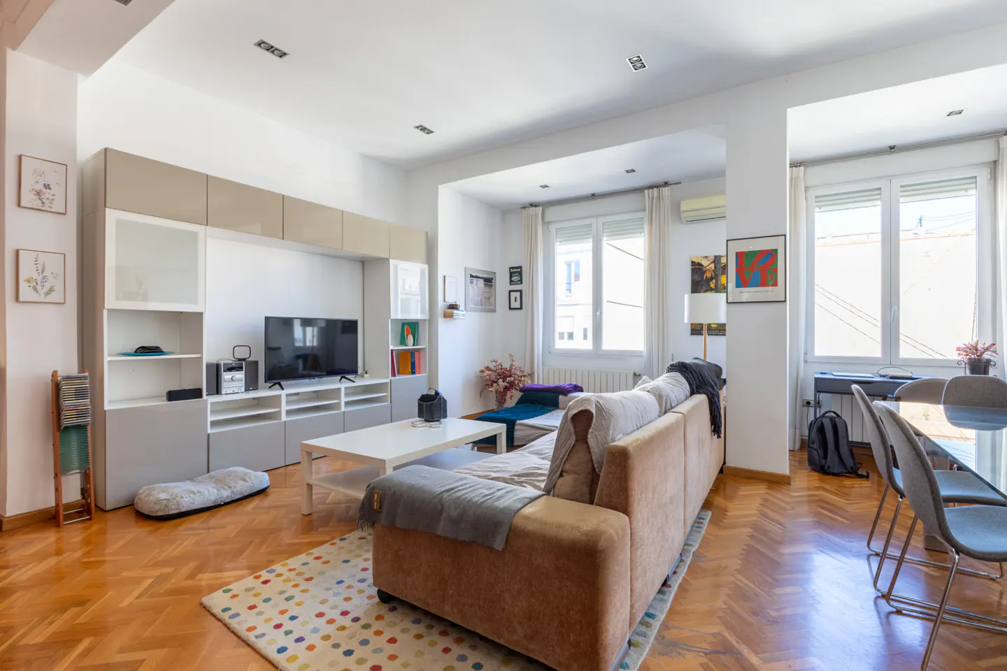 Bright living room with hardwood floors, a brown sofa, white walls, and a large entertainment center with a TV. Windows let in natural light.