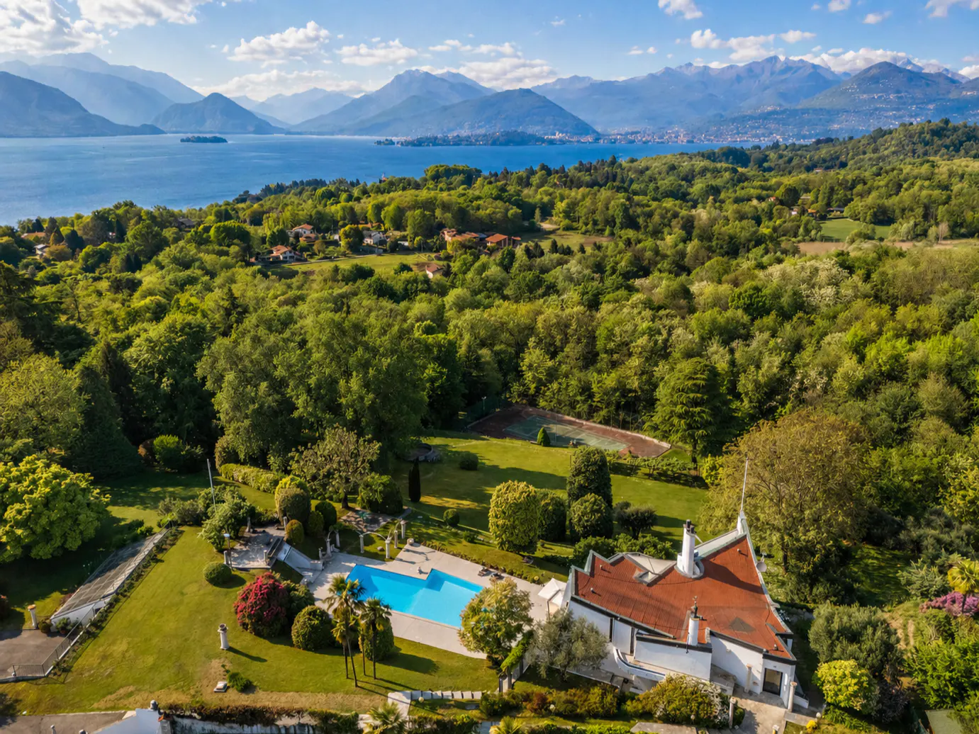 Aerial view of a white villa with a red roof, a blue pool, green lawns, and a lake with mountains in the background.