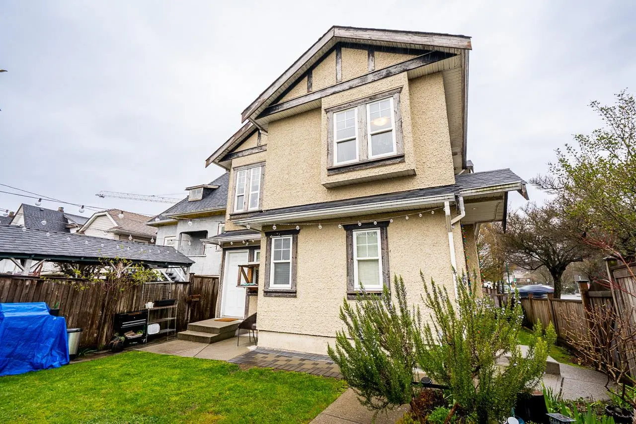 Two-story beige house with white-framed windows, a green lawn, and a wooden fence under a cloudy sky.