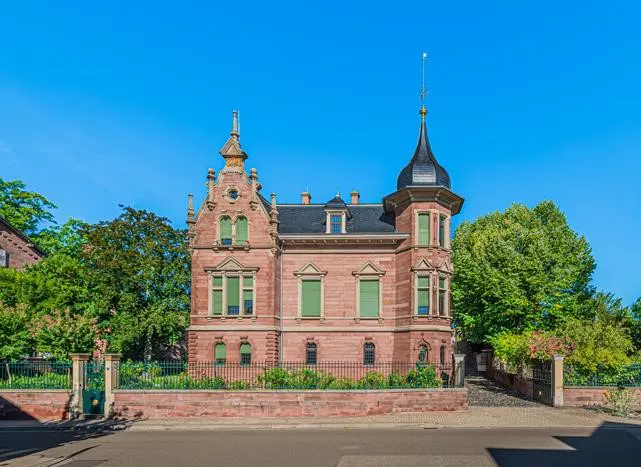 A three-story red brick house with green shutters and a black roof under a clear blue sky.