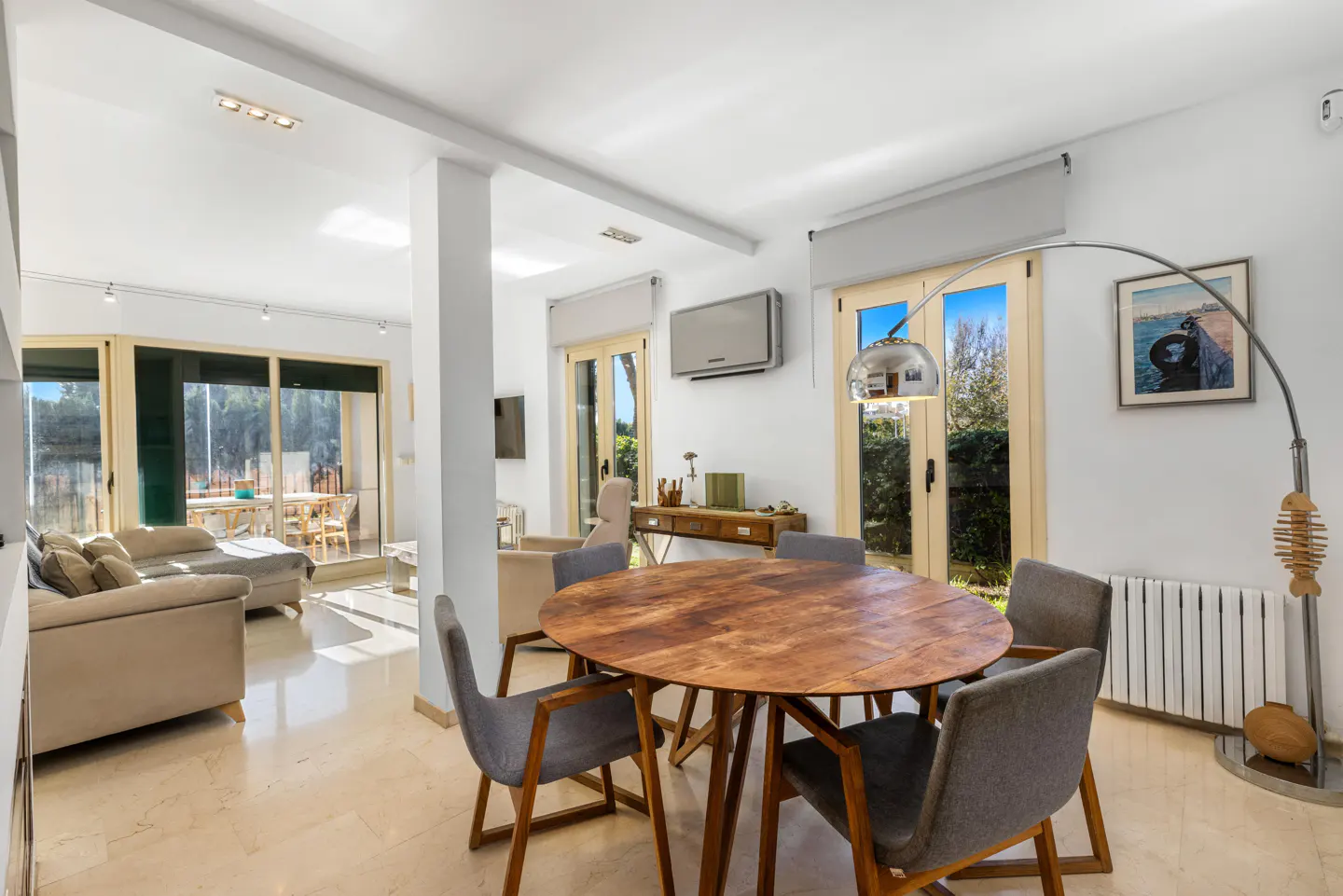 Bright, open-plan living and dining area with a round wooden table, gray chairs, and a beige sofa. Large windows offer outdoor views.