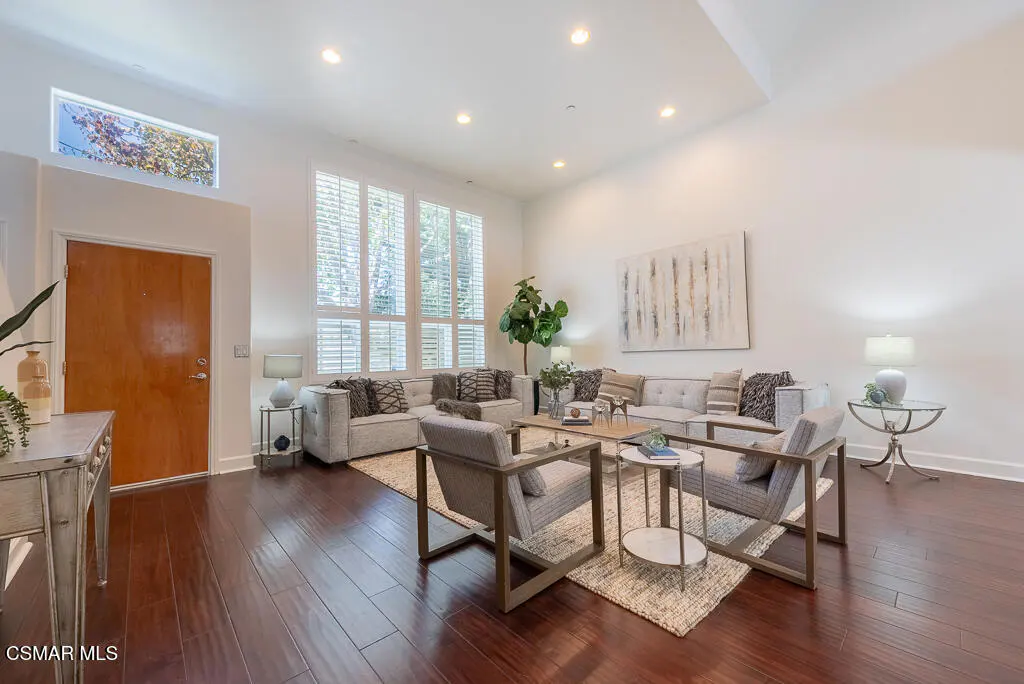 Bright living room with hardwood floors, white walls, and a large window. Gray sofas and chairs are arranged around a wooden coffee table.