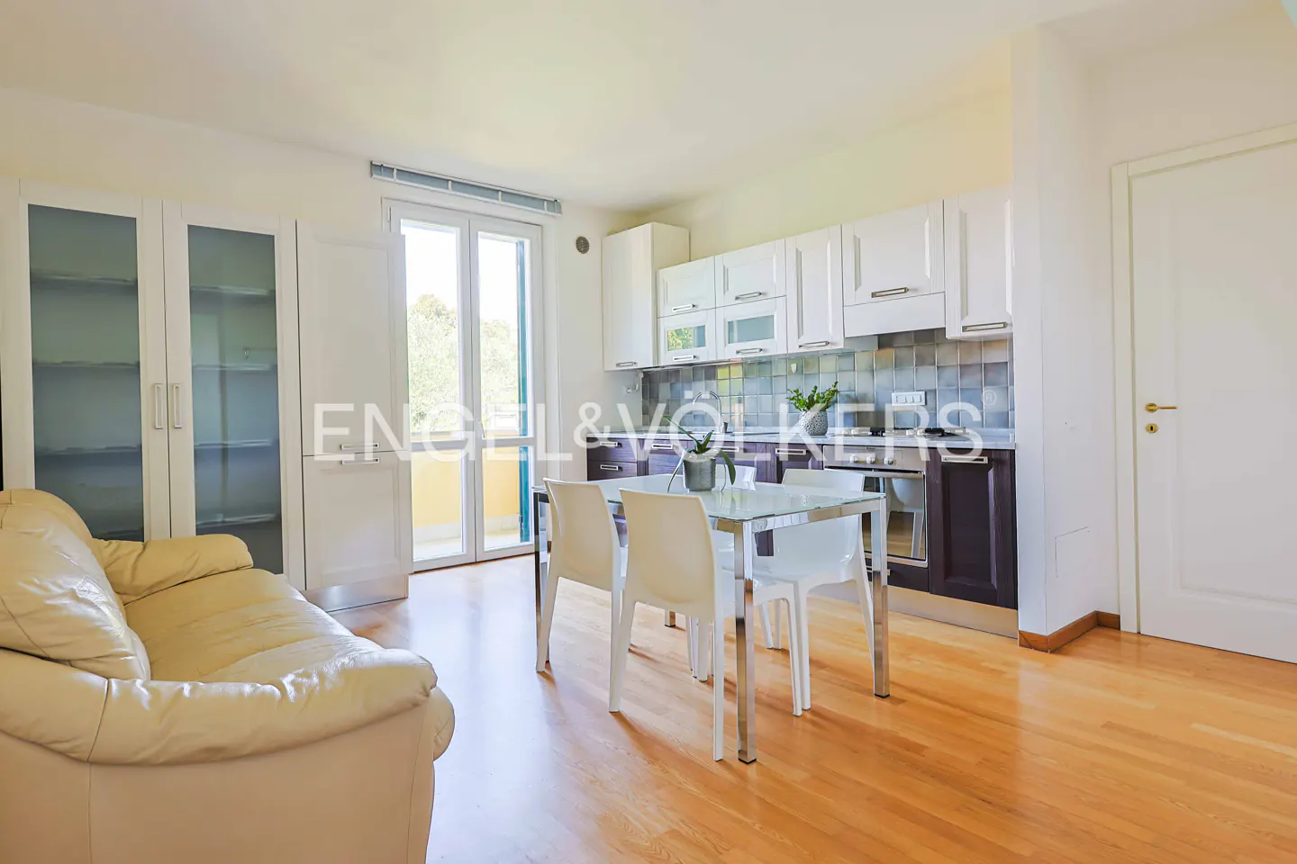 Bright apartment interior with hardwood floors, a cream-colored sofa, and a glass dining table with white chairs. White kitchen cabinets line the back wall.