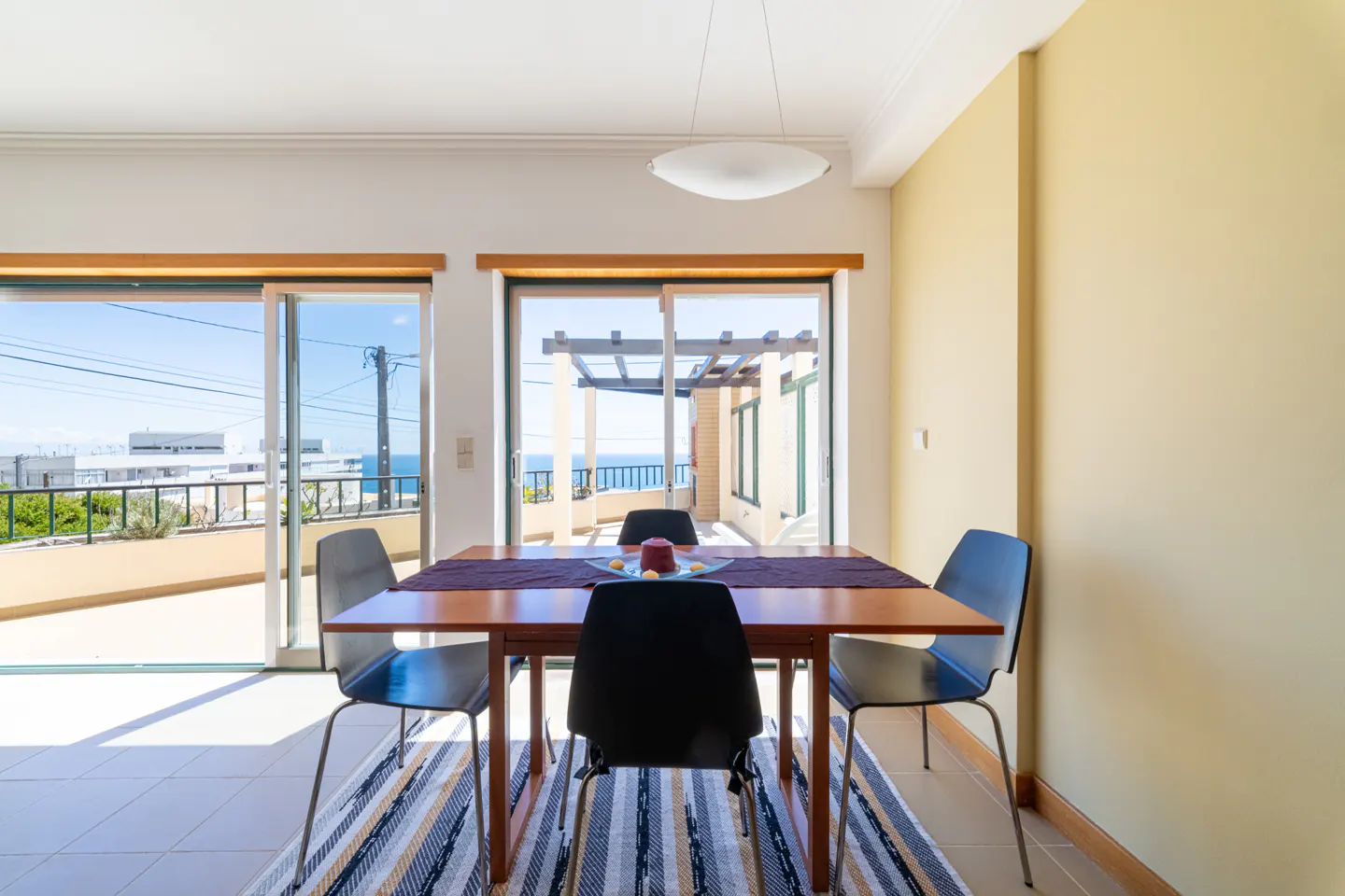 Dining room with a wooden table, four chairs, and a striped rug. Sliding glass doors open to a balcony with an ocean view.