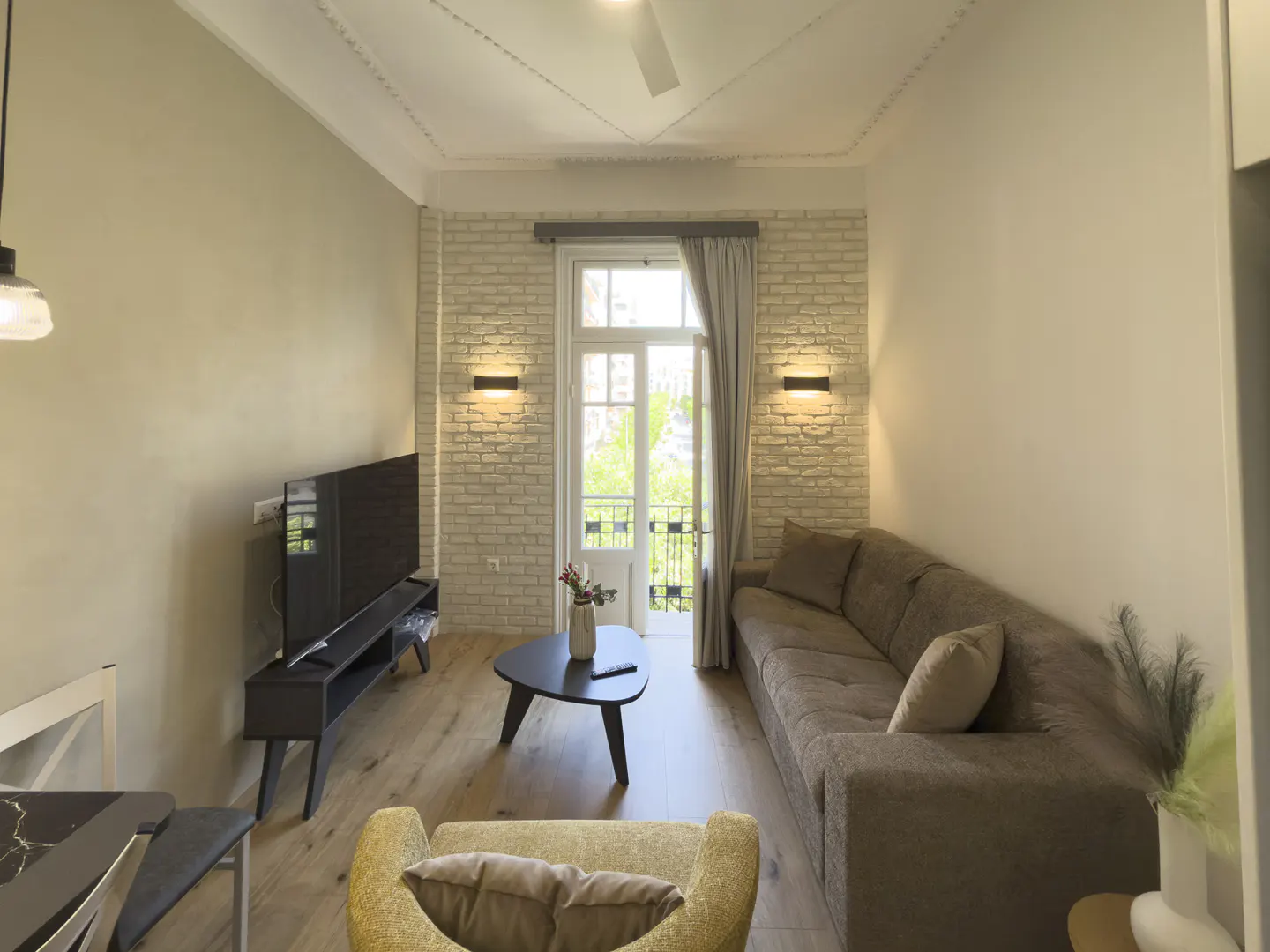 Living room with a brown sofa, yellow chair, and black TV. A white brick wall has sconces and a door to a balcony.