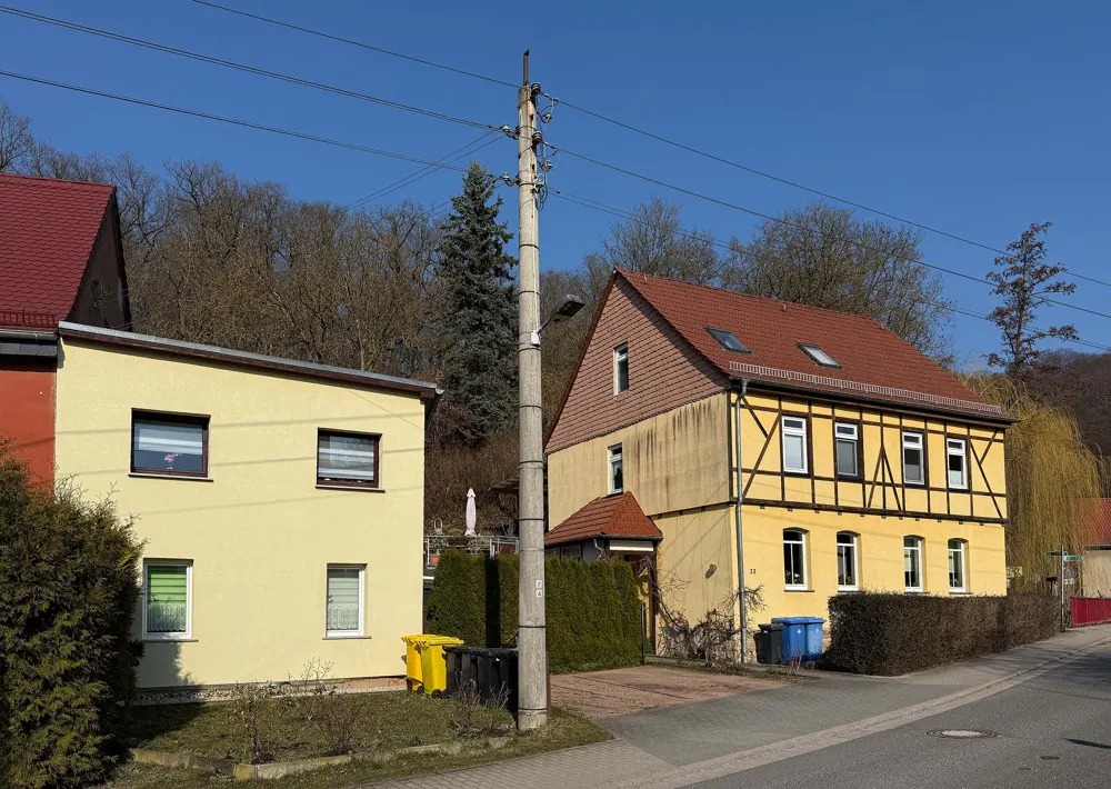 Two yellow houses, one with a red roof and timber framing, sit on a street under a clear blue sky. A utility pole stands in front.