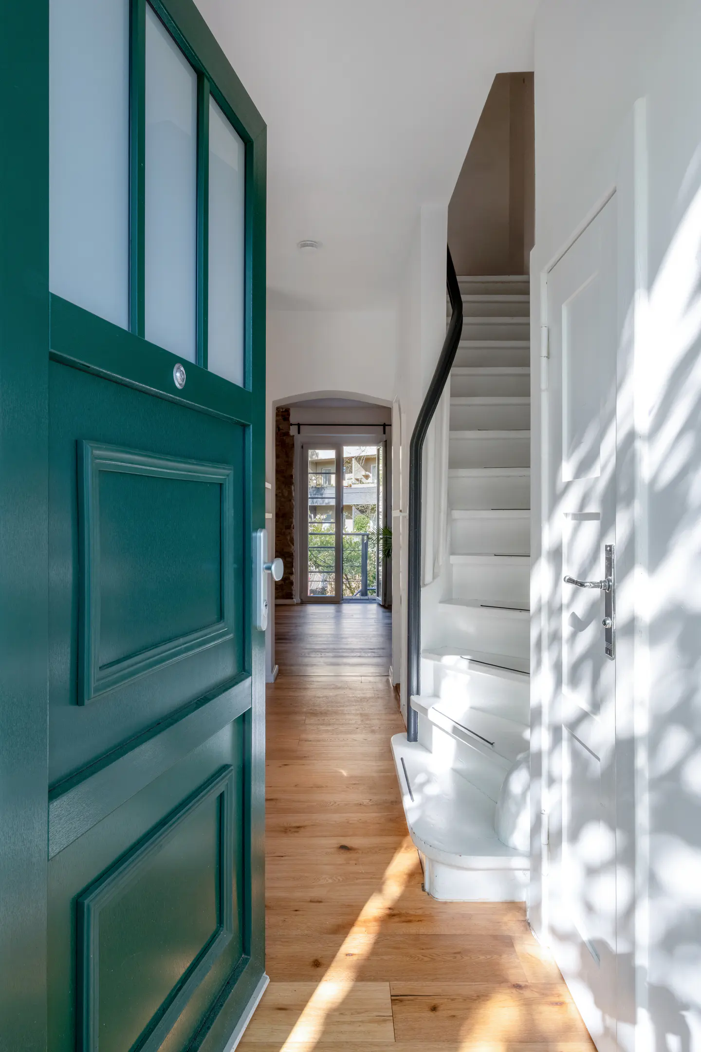 Open teal front door reveals a bright foyer with hardwood floors, white stairs with a black railing, and a glimpse of a sunlit room.
