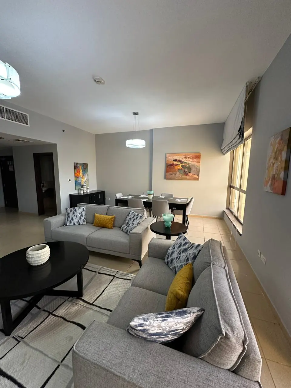 A modern living room with gray sofas, yellow pillows, and a black coffee table on a geometric rug. A dining table and artwork are in the background.