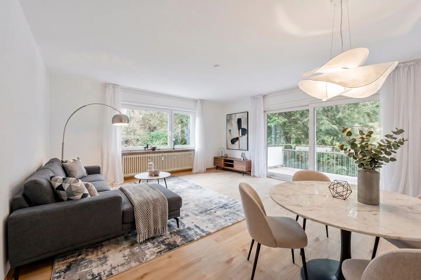 Bright living room with gray sofa, marble table, and modern light fixture. Windows overlook green trees.