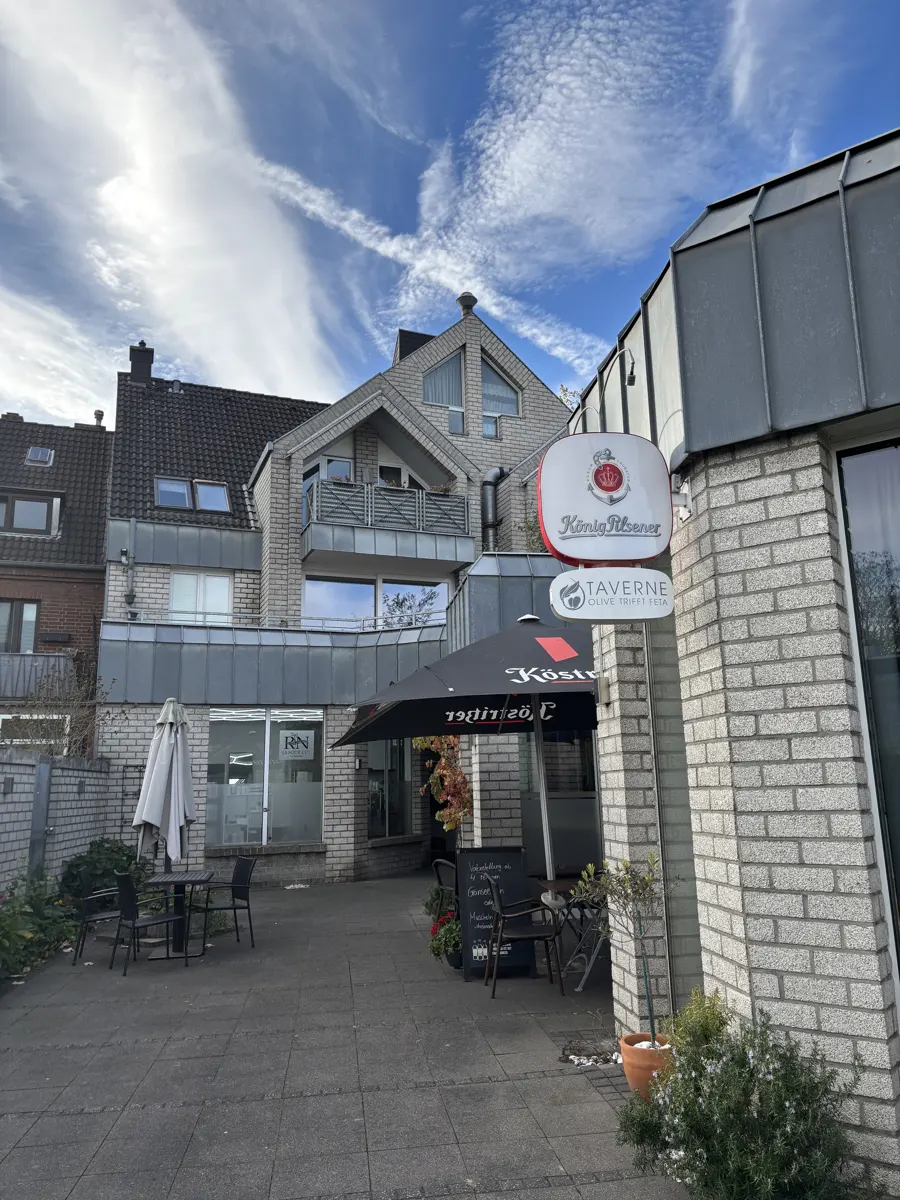 Exterior of a light brick building with "König Pilsener" and "Taverne" signs. Tables and chairs are on the patio.