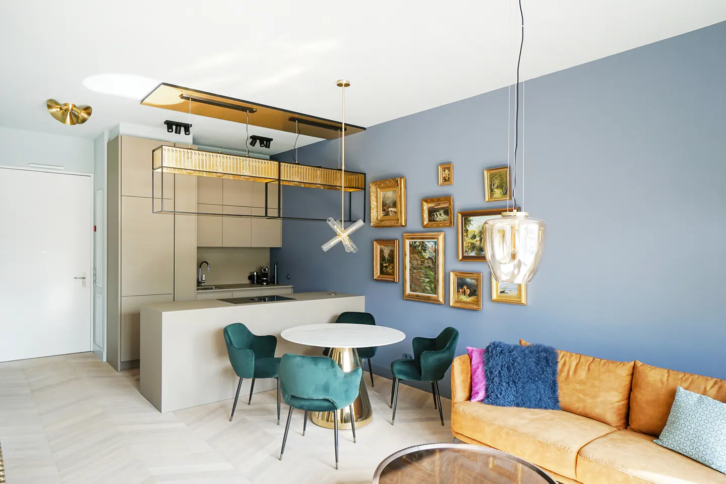 Interior view of a modern apartment with a kitchen, dining area, and living room. Blue wall with framed art, tan sofa, and green chairs.
