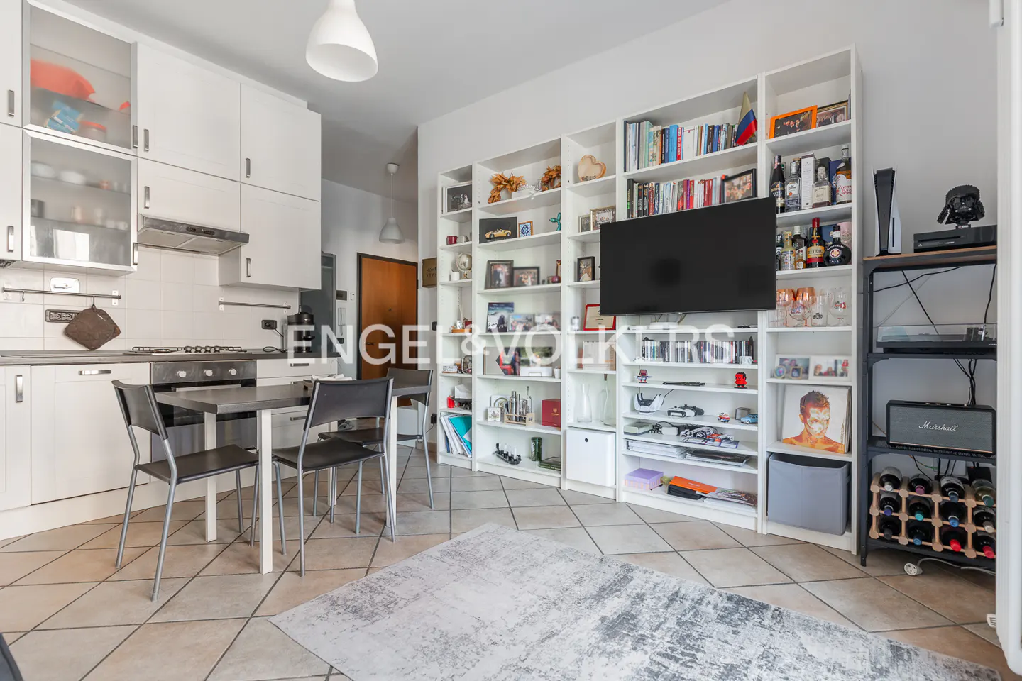 Bright living space with white kitchen cabinets, a dining table with black chairs, and a large white bookshelf with a TV.