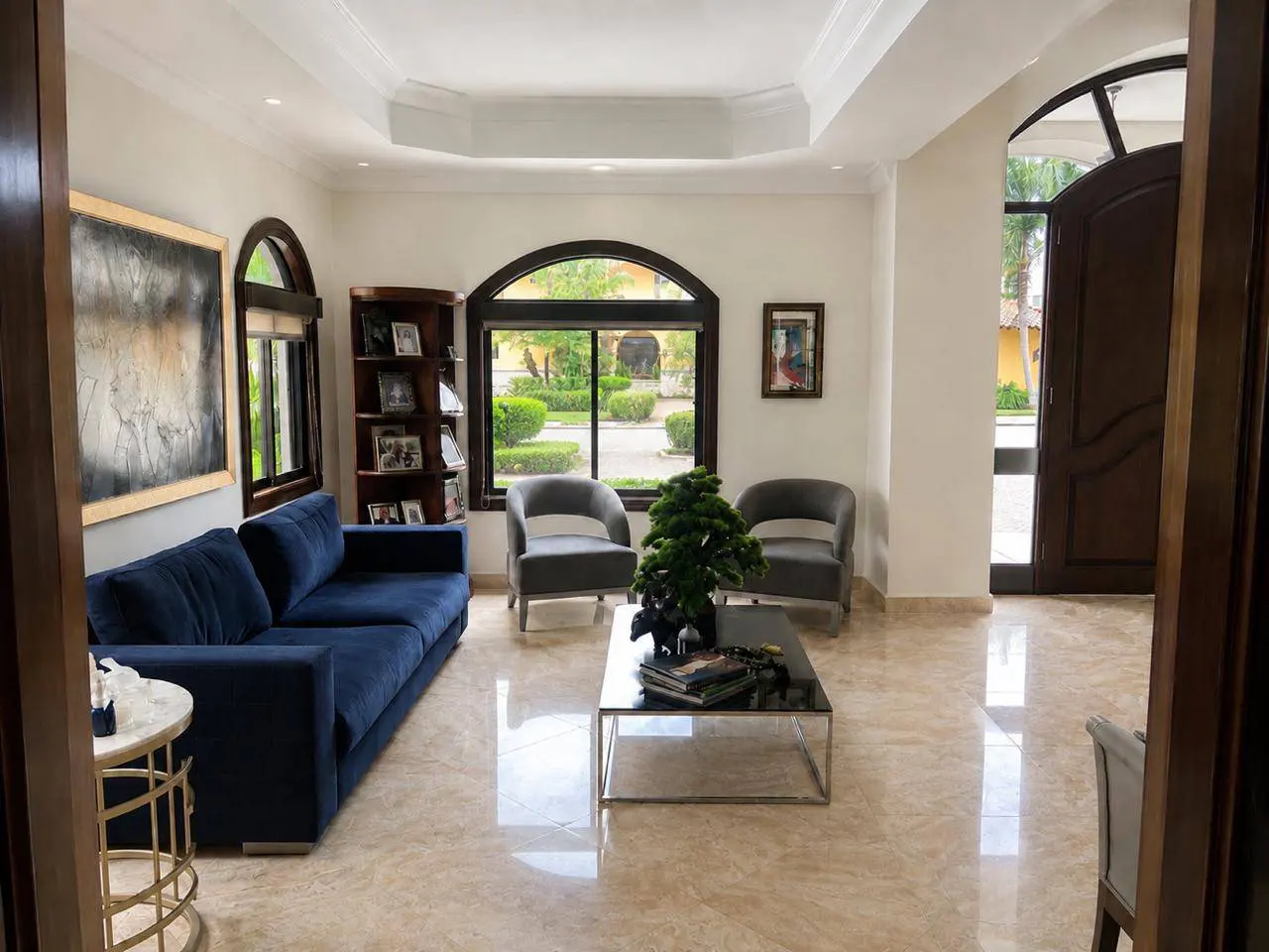 Bright living room with blue sofa, gray chairs, and a glass coffee table. Arched windows overlook a manicured garden.